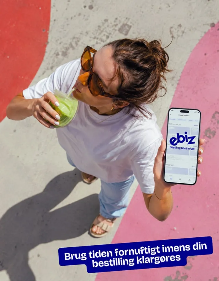 Kvinde med en grøn smoothie viser E-BIZ-appen på sin telefon, fotograferet oppefra. Tekst: Brug tiden fornuftigt imens din bestilling klargøres.
